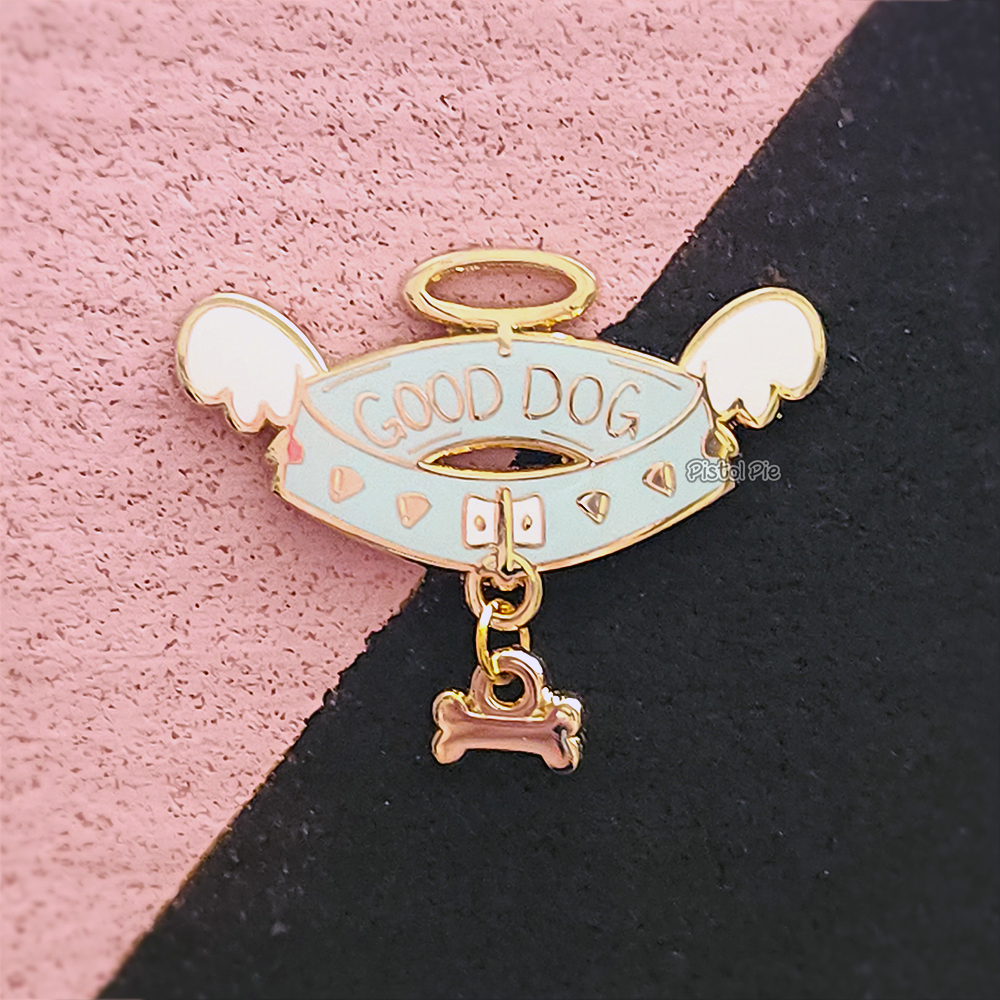 photo of an enamel pin by pistol pie of a spiked dog collar with dangling bone charm that has angel wings and text good dog