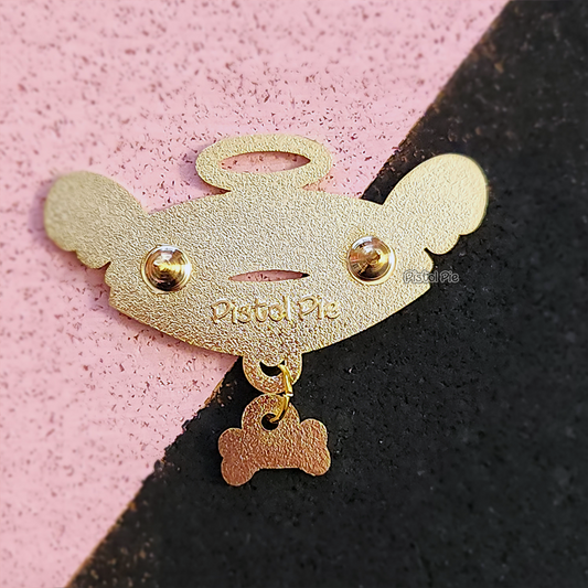 photo of the back of an enamel pin by pistol pie of a spiked dog collar with dangling bone charm that has angel wings and embedded Pistol Pie logo