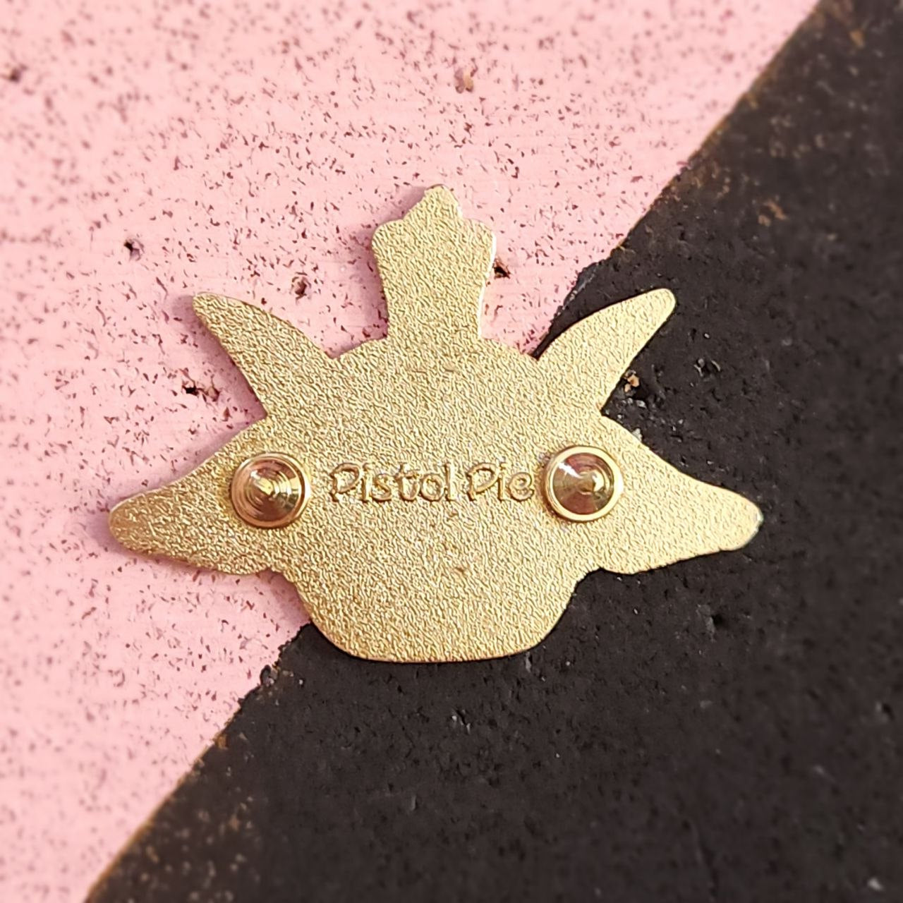 Gold enamel pin with 'Pistiol Pie' branding on a pink and black background