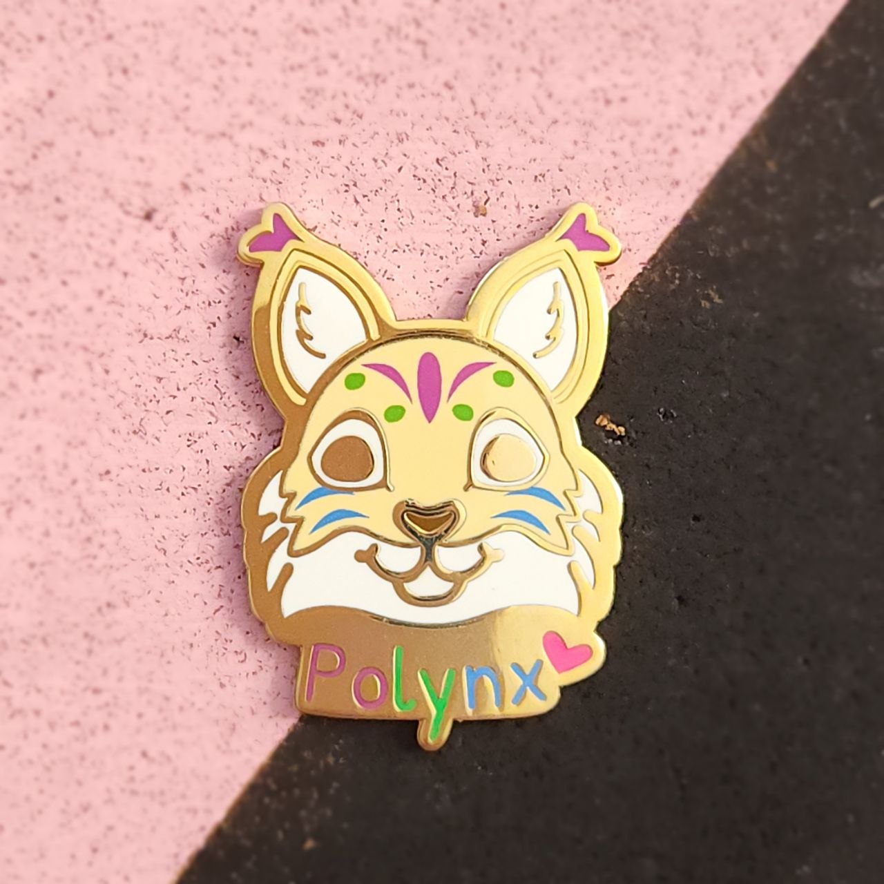 Enamel pin of a cartoon-like fface with 'Polynx' on a pink and black background
